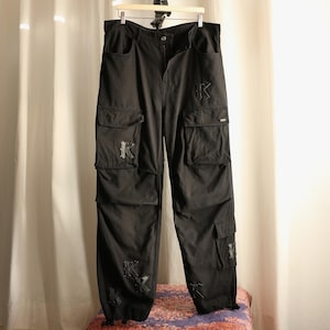 May include: Black cargo trousers featuring multiple utility pockets and several embroidered letter K patches in a gothic font. The trousers include a button and zip fly closure, belt loops, and adjustable drawstring hems at the bottom of each leg.
