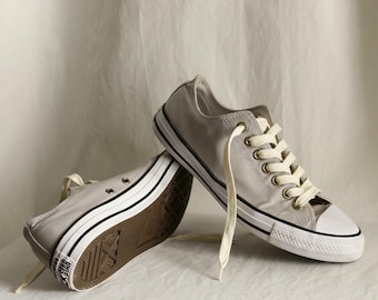 May include: Pair of light gray canvas sneakers with white laces and a white rubber toe cap. The sneakers have a black stripe along the white rubber sole and a brown rubber outsole. The shoes are a classic, casual style.