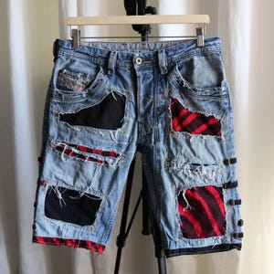 May include: Light blue denim shorts featuring a distressed finish with frayed edges and multiple patches. The patches consist of solid black fabric and red and black striped fabric. The shorts include side tabs and a button fly closure.