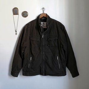 Könnte beinhalten: Dunkelbraune Jacke mit durchgehendem Reißverschluss, zwei Brusttaschen und zwei Seitentaschen. Die Jacke hängt an einem Holzbügel vor einer weißen Wand. Der Kragen hat den Text "C/N/R".