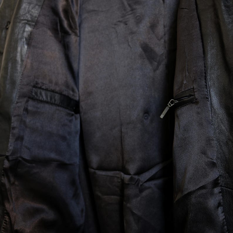May include: Close-up of the interior of a black leather jacket. The lining is a dark, satiny fabric with a pocket on the left side. A zipper pocket is visible on the right side. The jacket appears to be well-worn.
