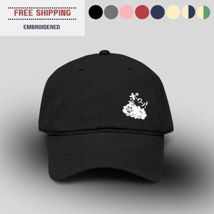 May include: A black baseball cap featuring a white embroidered graphic of a smiling cloud character with Japanese text above it. The cap has a structured crown, a curved brim, and a button on top, showcasing a clean and simple design.
