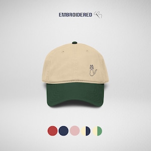 May include: A two-tone baseball cap with a beige crown and a dark green brim. The front features a small embroidered graphic of a cat wearing a bow tie. Above the cap, the text EMBROIDERED is displayed in dark blue, accompanied by a needle icon.