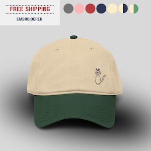 May include: A beige and dark green baseball cap with an embroidered cat design. The cap has a curved peak and a button on top. The text "EMBROIDERED" is visible on the top left.