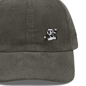 May include: A dark olive green corduroy baseball cap featuring a small, white, pixelated embroidered graphic of a character on the front panel. The cap has a structured crown, a curved brim, and visible vertical corduroy texture throughout.