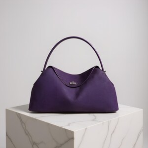 May include: A deep purple handbag with a curved handle and a silver-toned clasp. The bag has a structured shape and sits on a white marble block. The bag's design is simple and elegant.