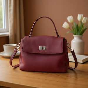 May include: A burgundy leather handbag with a top handle and a detachable shoulder strap. The bag features a flap closure with a silver-tone clasp. The bag is sitting on a wooden table next to a cup of coffee and a vase of white flowers.