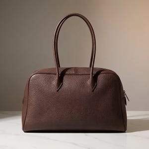 Könnte beinhalten: Eine dunkelbraune Handtasche mit genarbter Lederstruktur, ausgestattet mit zwei hohen, schmalen Tragegriffen und einem horizontalen Reißverschluss an der Oberseite. Die Tasche hat eine abgerundete, strukturierte rechteckige Form.