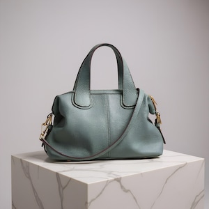 May include: A light teal leather handbag with a top handle and a detachable shoulder strap. The bag has gold-colored hardware and is displayed on a white marble block. The bag has a structured shape.