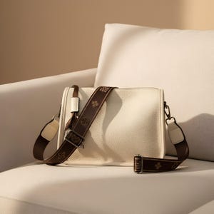 May include: Cream-colored crossbody bag with a textured finish. The bag features a brown patterned strap and a smaller matching strap. The bag is resting on a white sofa.