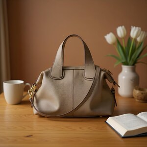 May include: A beige leather handbag with a top handle and a detachable shoulder strap sits on a wooden table. The bag has a rounded shape and gold-colored hardware. A white mug, a vase of white tulips, and an open book are also on the table.