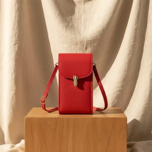 May include: A red leather crossbody bag with a flap closure and a gold-colored clasp. The bag has a long, adjustable strap and is displayed on a wooden block against a neutral fabric backdrop.