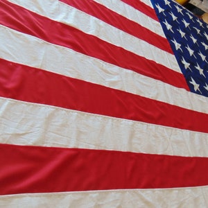 Può includere: Grande bandiera americana con strisce rosse e bianche e un cantone blu pieno di stelle bianche. La bandiera è in tessuto e viene mostrata per intero, mostrando le sue dimensioni. Simbolo degli Stati Uniti.