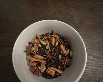 May include: A white ceramic bowl containing a mix of tea leaves, cinnamon sticks, star anise, cardamom pods, and other spices. The tea blend is dark brown with green and tan highlights. The bowl rests on a dark wooden surface.