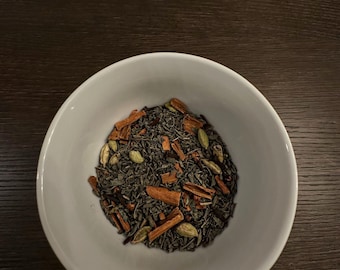 May include: A white ceramic bowl containing loose leaf tea, cinnamon sticks, and green cardamom pods. The tea leaves are dark green, and the cinnamon sticks are brown. The bowl is on a dark brown wooden surface.