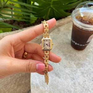 Puede incluir: Un reloj de pulsera dorado con una esfera rectangular y números romanos. El reloj tiene una delicada correa de eslabones. El reloj se sostiene sobre un fondo de follaje verde y una bebida en un vaso transparente.