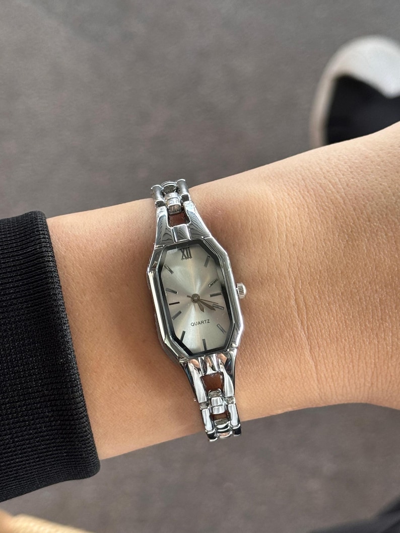 Pu&ograve; includere: Un orologio al quarzo color argento con quadrante rettangolare e cinturino argentato. Il quadrante dell'orologio &egrave; di colore chiaro con lancette nere e numeri romani. L'orologio &egrave; indossato al polso.