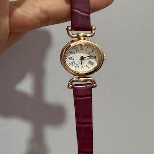 Peut inclure: Une montre à quartz dorée avec un cadran blanc et des chiffres romains noirs. La montre a un bracelet en cuir bordeaux avec une texture crocodile. Le cadran de la montre est de forme ovale.