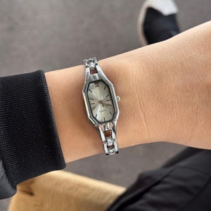 Pu&ograve; includere: Un orologio al quarzo color argento con un quadrante ottagonale allungato e un quadrante di colore chiaro. L'orologio presenta un bracciale a maglie e indici delle ore con numeri romani. L'orologio &egrave; lungo circa 2,5 cm.