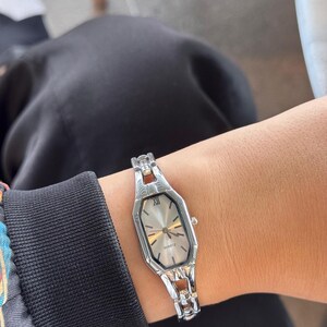 Pu&ograve; includere: Orologio al quarzo color argento con quadrante rettangolare e cinturino argentato. L'orologio ha un quadrante di colore chiaro con lancette e indici dorati. L'orologio &egrave; indossato al polso.
