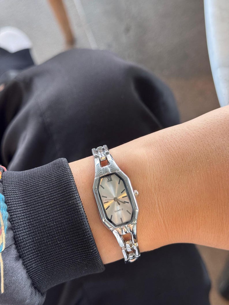 Pu&ograve; includere: Orologio al quarzo color argento con quadrante rettangolare e cinturino argentato. L'orologio ha un quadrante di colore chiaro con lancette e indici dorati. L'orologio &egrave; indossato al polso.