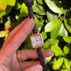 Puede incluir: Un reloj de cuarzo cuadrado dorado con esfera blanca de nácar e indicadores de hora con números romanos. El reloj tiene una correa de cuero marrón oscuro texturizado. El reloj mide aproximadamente 1,9 cm de ancho.