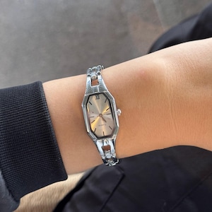Pu&ograve; includere: Orologio al quarzo color argento con quadrante ottagonale e cinturino argentato. L'orologio ha un quadrante chiaro con lancette argentate e numeri romani. L'orologio &egrave; indossato su un polso, con una manica nera e abiti neri sullo sfondo.