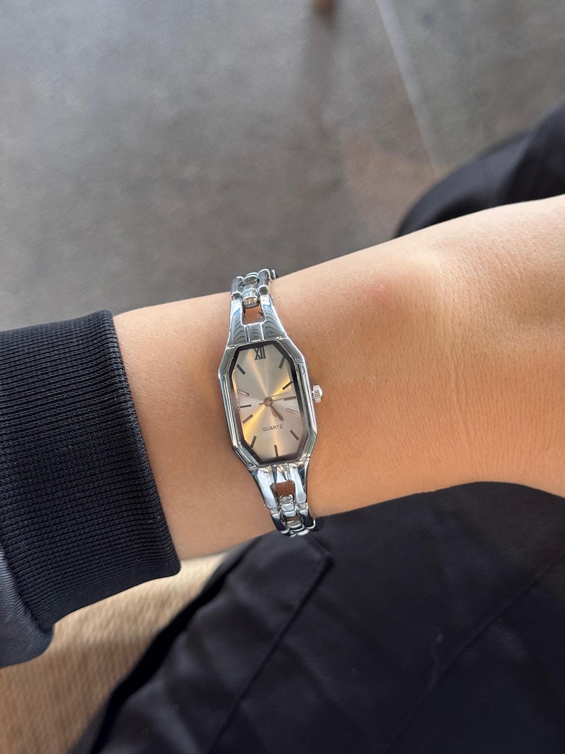 Pu&ograve; includere: Orologio al quarzo color argento con quadrante ottagonale e cinturino argentato. L'orologio ha un quadrante chiaro con lancette argentate e numeri romani. L'orologio &egrave; indossato su un polso, con una manica nera e abiti neri sullo sfondo.