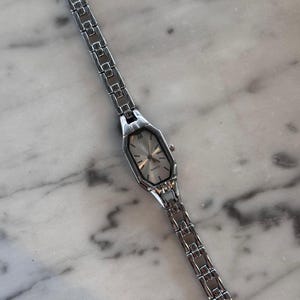 Pu&ograve; includere: Un orologio da polso argentato con quadrante rettangolare e cinturino a maglie. Il quadrante &egrave; argentato con lancette e indici neri. La parola "QUARTZ" &egrave; stampata sul quadrante. L'orologio &egrave; su una superficie di marmo.