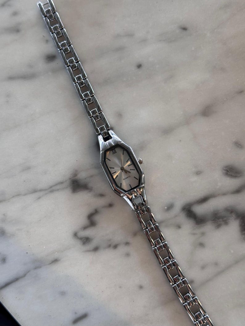 Pu&ograve; includere: Un orologio da polso argentato con quadrante rettangolare e cinturino a maglie. Il quadrante &egrave; argentato con lancette e indici neri. La parola "QUARTZ" &egrave; stampata sul quadrante. L'orologio &egrave; su una superficie di marmo.