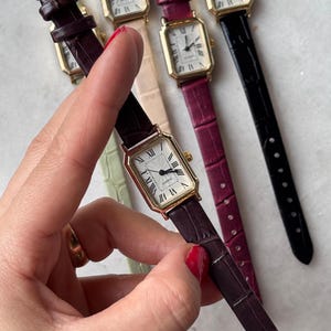 Könnte beinhalten: Eine Sammlung rechteckiger Quarzuhren mit goldfarbenen Gehäusen und Lederarmbändern in verschiedenen Farben, darunter Burgunderrot, Schwarz und Hellgrün. Die Zifferblätter haben römische Ziffern und schwarze Zeiger.