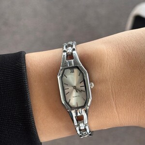 Pu&ograve; includere: Orologio al quarzo color argento con quadrante rettangolare e cinturino a maglie. Il quadrante ha uno sfondo argentato con lancette e indici neri. L'orologio &egrave; su un polso.