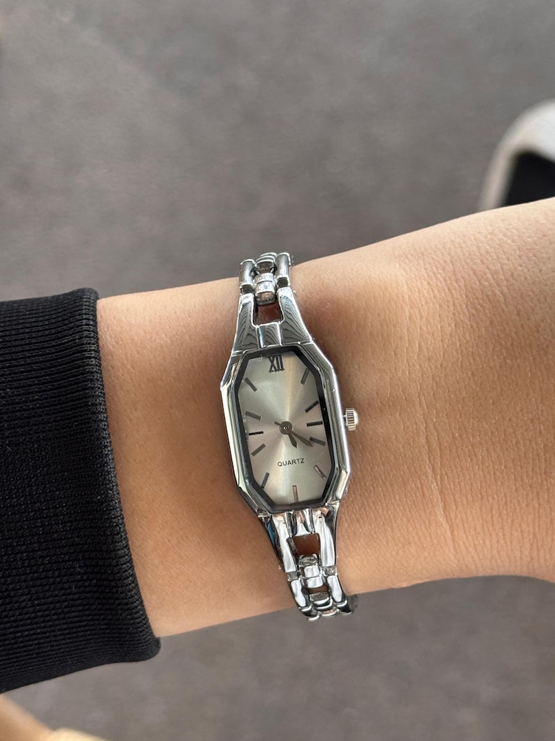 Pu&ograve; includere: Orologio al quarzo color argento con quadrante rettangolare e cinturino a maglie. Il quadrante ha uno sfondo argentato con lancette e indici neri. L'orologio &egrave; su un polso.