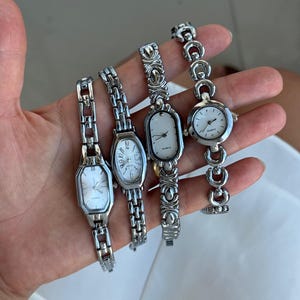 May include: Four silver-toned quartz wristwatches with metal link bands. The watches feature various case shapes including octagonal, oval, and round designs, each with a white dial, silver hands, and the word Quartz printed on the watch face.