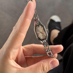Pu&ograve; includere: Un orologio al quarzo argentato con quadrante rettangolare e bracciale a maglie. Il quadrante dell'orologio &egrave; di colore chiaro con lancette argentate e numeri romani. L'orologio &egrave; tenuto davanti a uno sfondo grigio.