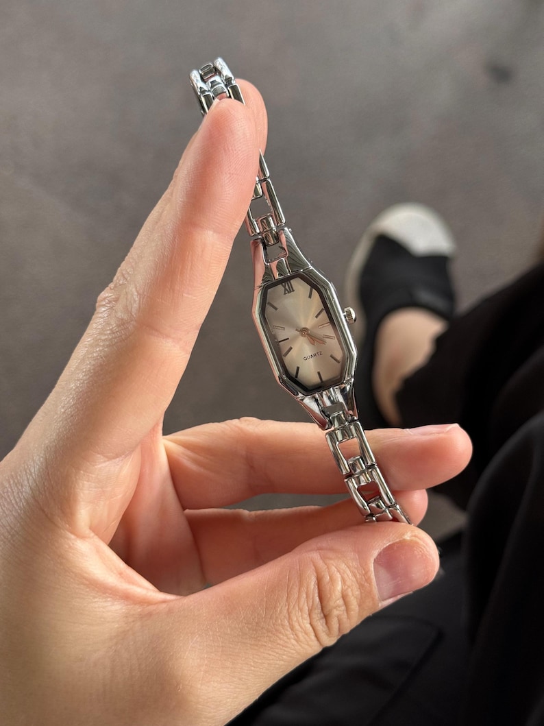 Pu&ograve; includere: Un orologio al quarzo argentato con quadrante rettangolare e bracciale a maglie. Il quadrante dell'orologio &egrave; di colore chiaro con lancette argentate e numeri romani. L'orologio &egrave; tenuto davanti a uno sfondo grigio.