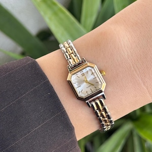 May include: A gold and silver quartz watch with a rectangular face and a silver band. The watch face is a light colour with gold Roman numerals. The watch is on a wrist.