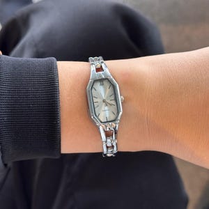 Pu&ograve; includere: Orologio al quarzo argentato con quadrante rettangolare e cinturino argentato. L'orologio ha un quadrante chiaro con lancette nere e numeri romani. Il cinturino ha dettagli argentati e marroni. L'orologio &egrave; indossato al polso.
