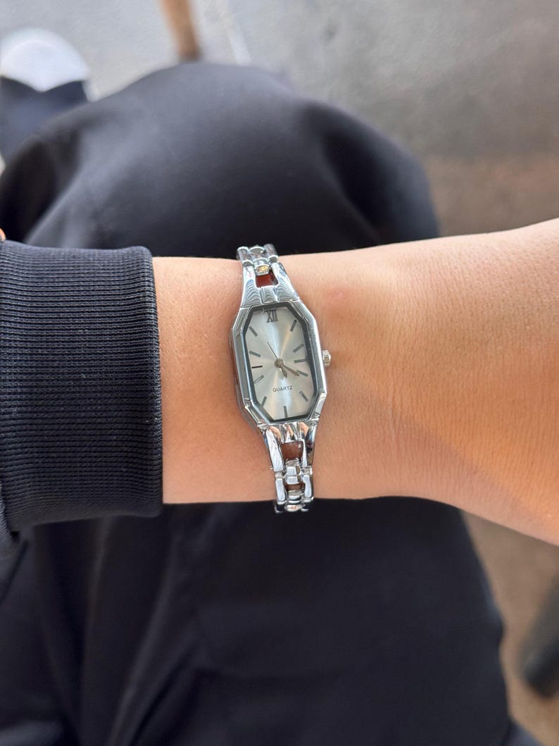 Pu&ograve; includere: Orologio al quarzo argentato con quadrante rettangolare e cinturino argentato. L'orologio ha un quadrante chiaro con lancette nere e numeri romani. Il cinturino ha dettagli argentati e marroni. L'orologio &egrave; indossato al polso.