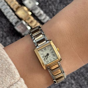 Könnte beinhalten: Eine rechteckige Quarzuhr mit goldfarbener Lünette und einem silber- und goldfarbenen Armband. Das Zifferblatt der Uhr ist weiß mit schwarzen römischen Ziffern und Zeigern. Weitere Uhren sind im Hintergrund zu sehen.