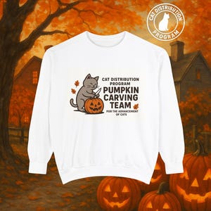以下が含まれることがあります： 白いスウェットシャツ。灰色の猫がカボチャを彫っているグラフィック。テキストは「Cat Distribution Program Pumpkin Carving Team for the Advancement of Cats」と書かれています。円形のロゴには「Cat Distribution Program」と書かれています。