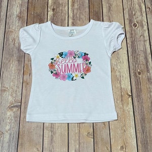Girls white puff short sleeve tee shirt with the Hello Summer floral design on the front.  The tee shirt is laying flat on the brown wooden floor.