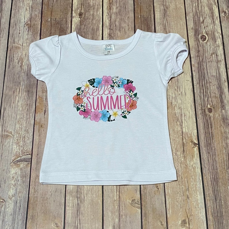 Girls white puff short sleeve tee shirt with the Hello Summer floral design on the front.  The tee shirt is laying flat on the brown wooden floor.