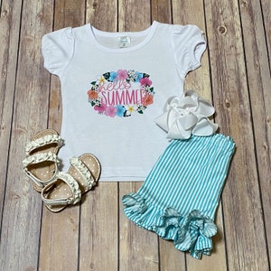 Girls white tee decorated with the Hello Summer floral design  paired with a turquoise and white striped shorts set laying flat.  A pair of sandals and a white hair bow have been added to the layout.  The background is a brown wood flooring.