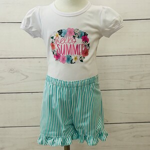 Girls tee and short set on a mannequin with a white background. The tee is white and has puff short sleeves with a Hello Summer floral design.  The shorts are turquoise and  white striped with an elastic waistband and ruffled legs hems