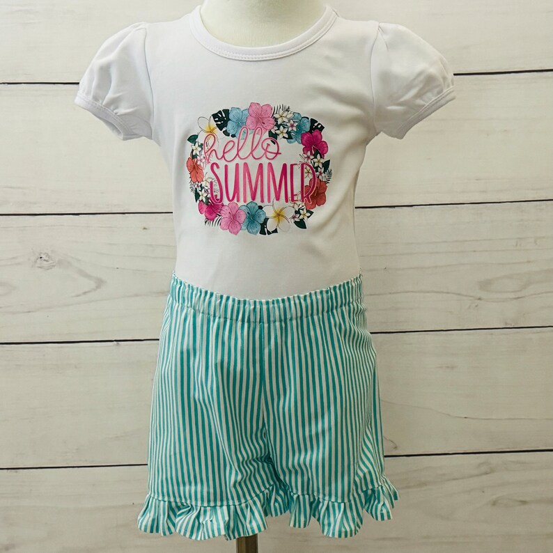 Girls tee and short set on a mannequin with a white background. The tee is white and has puff short sleeves with a Hello Summer floral design.  The shorts are turquoise and  white striped with an elastic waistband and ruffled legs hems