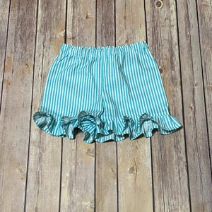 Girls turquoise and white striped shorts has an elstic waistband and ruffled leg hems. 
The shorts are laying flat on the brown wooden floor.
