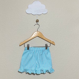 The turquoise and white striped shorts clipped to a hanger that is hooked over a wall clothes hook shaped like a cloud with a beige wall as the backgorund.