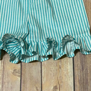 A close up view of the truquoise and white striped shorts ruffled leg hems against the brown flooring.