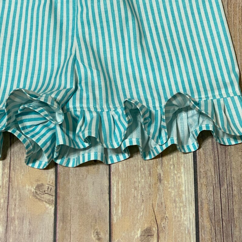 A close up view of the truquoise and white striped shorts ruffled leg hems against the brown flooring.